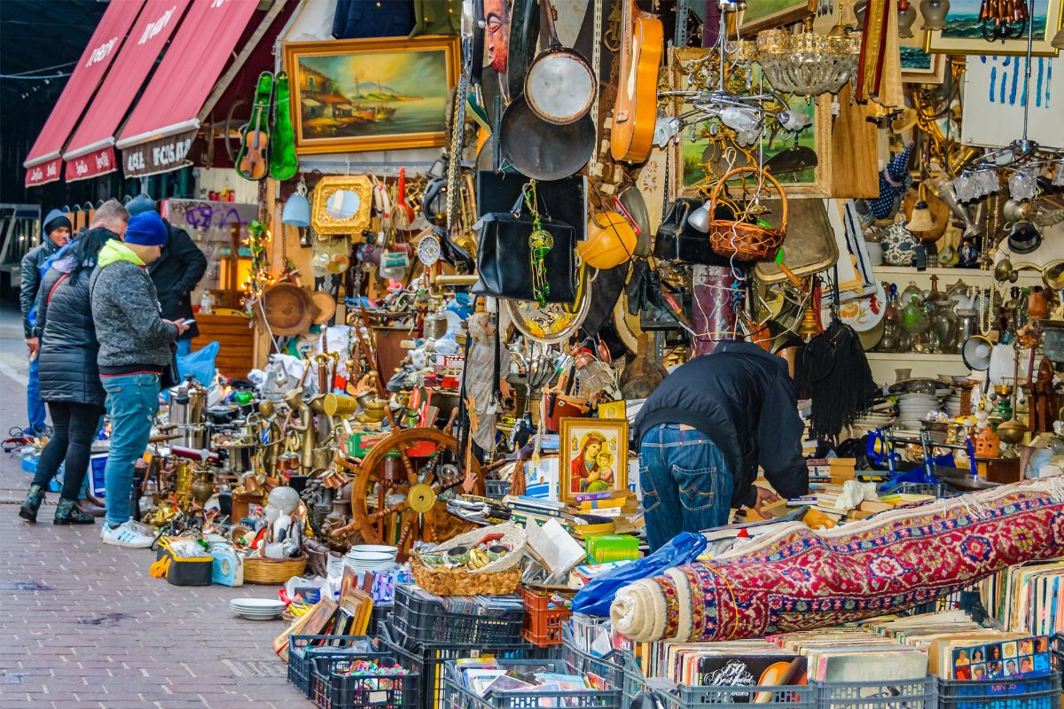 A bustling scene at Monastiraki Flea Market in Athens, showcasing people browsing through antique items, vintage goods, and various second-hand treasures, including rugs, paintings, and brassware.